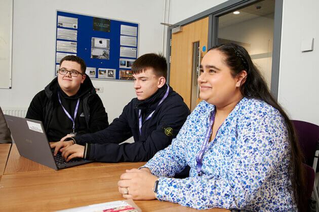 Three adult learners sat at a desk with a laptop in front of them