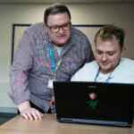 A student sat at a table looking at a laptop with a lecturer stood behind also looking at the laptop