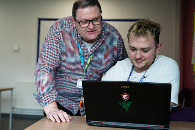 A student sat at a table looking at a laptop with a lecturer stood behind also looking at the laptop