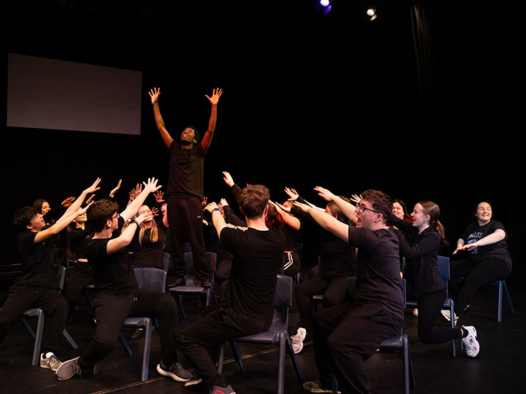 A group of students taking part in an acting performance sat on chairs with their hands in the air