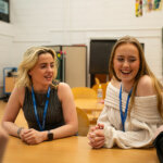 Two smiling students sat at a desk chatting to friends