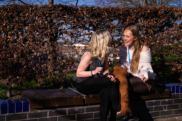 Two smiling students chatting sat on a wall with bushes behind them
