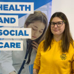 A female in a yellow long sleeved top stood in front of a banner that says 'Health and Social Care'