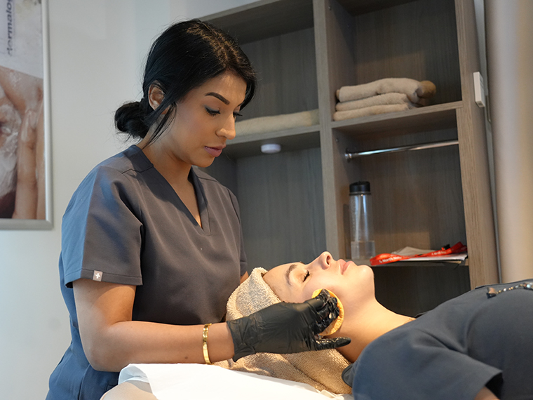 A lady with black hair, grey uniform and black gloves with her hands on a clients face who's laying on a treatment bed