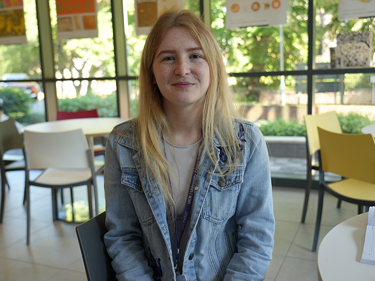 A female with long blonde hair and a light blue denim jacket sat at a table, a wall of windows in the background