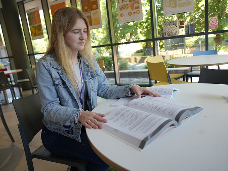 A female with long blonde hair and a light blue denim jacket sat at a table looking down at an open book, a wall of windows in the background
