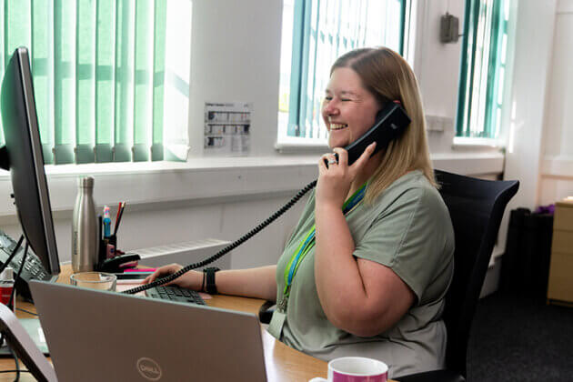 A smiling female sat at a desk looking towards a computer screen whilst on the phone