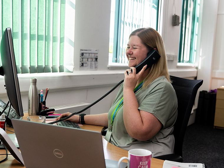 A smiling female sat at a desk looking towards a computer screen whilst on the phone