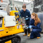 Two students in overalls bent down looking at machinery