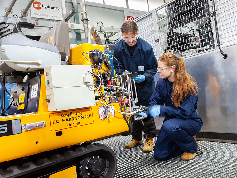 Two students in overalls bent down looking at machinery