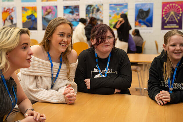 Four smiling students sat together chatting and leaning on a table, artwork up on the wall in the background