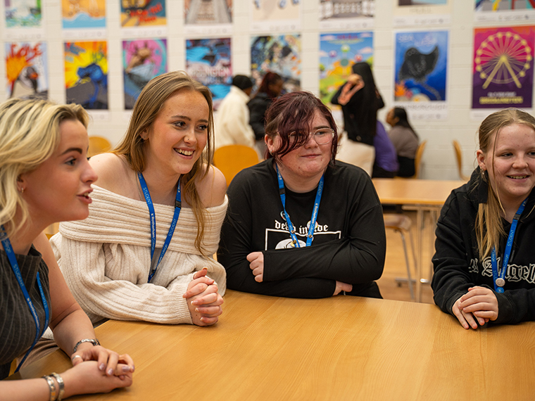 Four smiling students sat together chatting and leaning on a table, artwork up on the wall in the background
