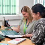 Two ladies sat at a desk with a computer in front of them, both looking down at a paper brochure