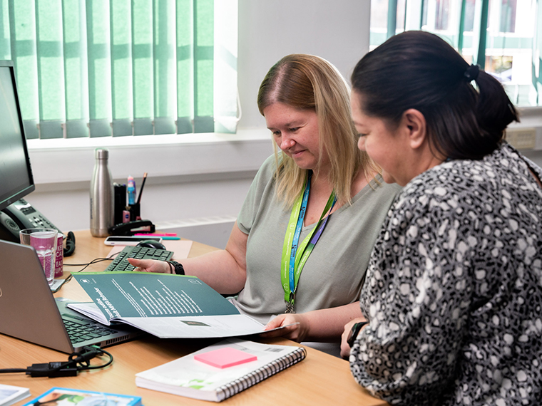Two ladies sat at a desk with a computer in front of them, both looking down at a paper brochure