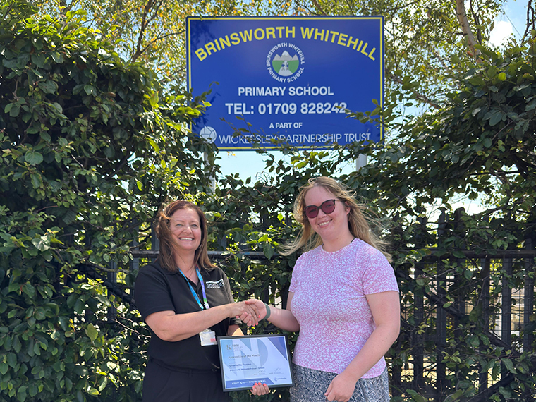 Two females stood shaking hands in front of a sign for Brinsworth Whitehall Primary School