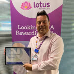 A smiling male holding a framed certificate, a pull up banner behind him that says Lotus