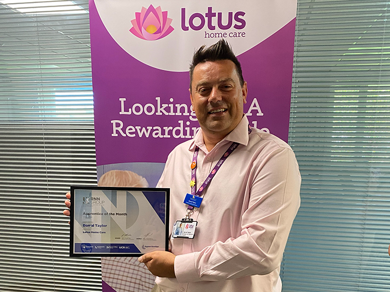 A smiling male holding a framed certificate, a pull up banner behind him that says Lotus