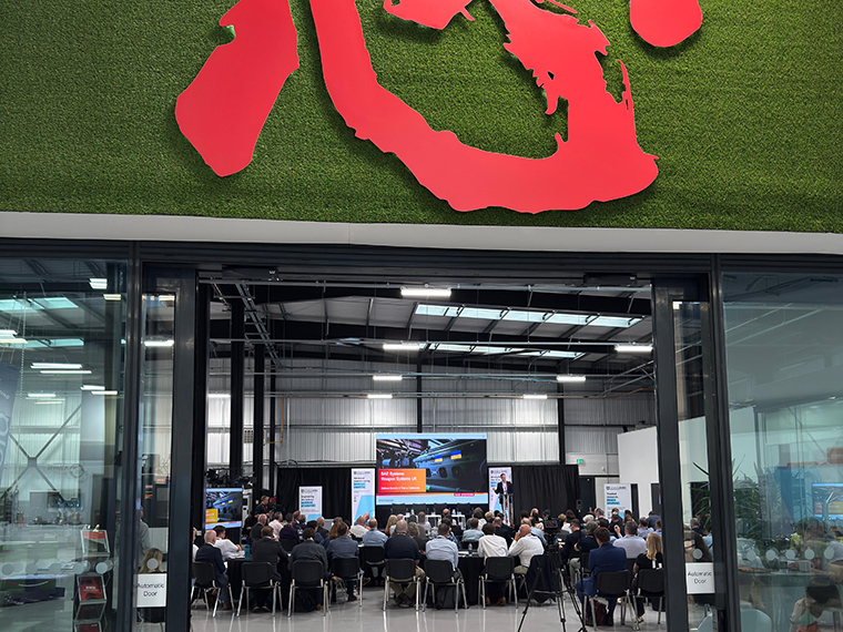 Looking through the open glass doors inside a building at a seated audience looking at a stage with a presentation screen on