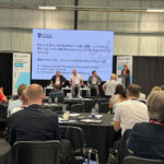 Four people on a stage with a presentation screen behind them, people sat in the audience watching on