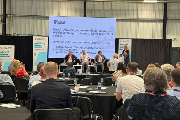 Four people on a stage with a presentation screen behind them, people sat in the audience watching on
