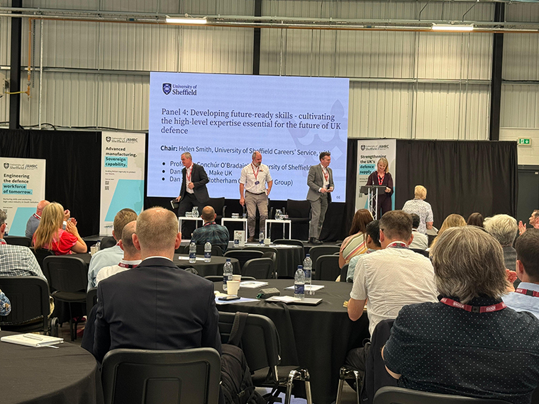 Four people on a stage with a presentation screen behind them, people sat in the audience watching on