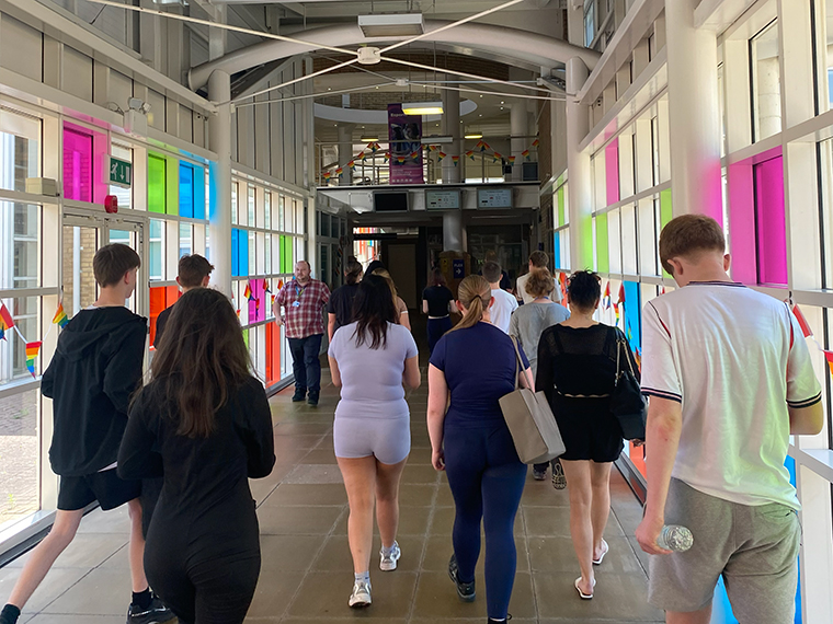 The back of a small crowd of people walking down an indoor corridor with colourful windows either side of them