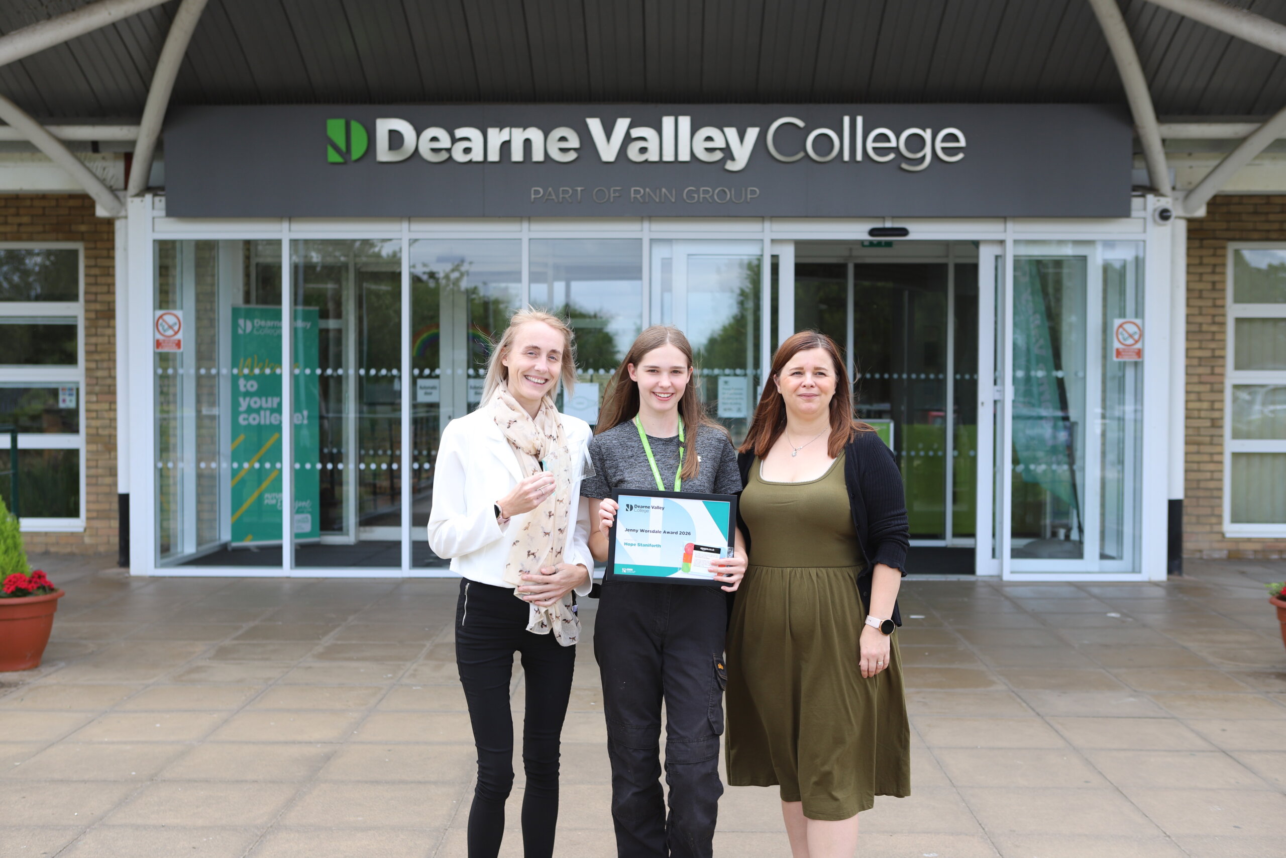 Three females stood together outside the Dearne Valley College building, the person in the centre holding a certificate