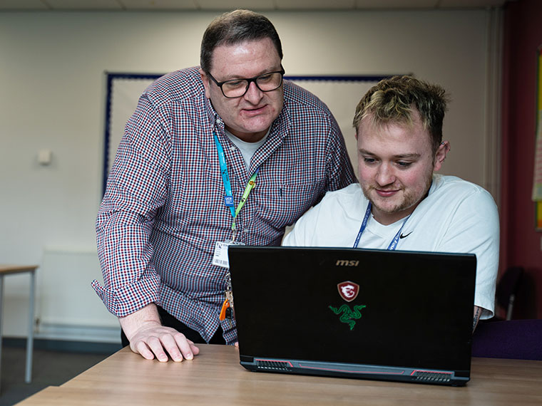A student sat at a table looking at a laptop with a lecturer stood behind also looking at the laptop