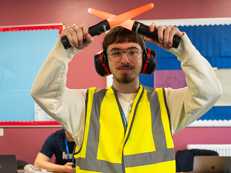 A male from the waist up wearing a high vis jacket and ear protectors holding an illuminated wand in each hand crossing them over his head