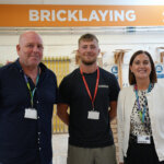 Two males and a female stood together, an orange and white sign above them that says 'Bricklaying'