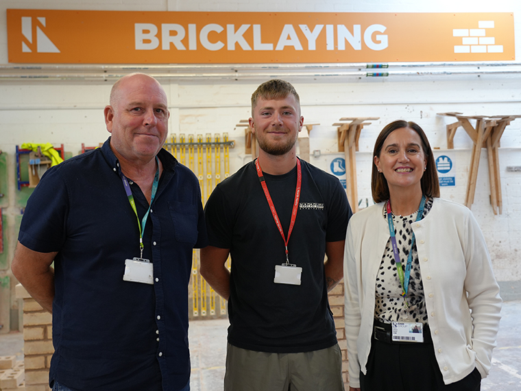 Two males and a female stood together, an orange and white sign above them that says 'Bricklaying'
