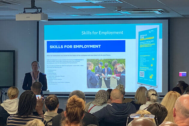 A female stood in front of a seated audience next to a large screen with 'Skills For Employment' written on it