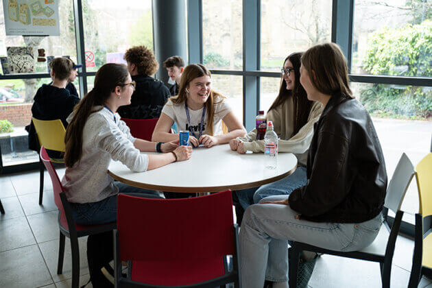 Two tables each full of smiling students socialising, large windows behind the tables