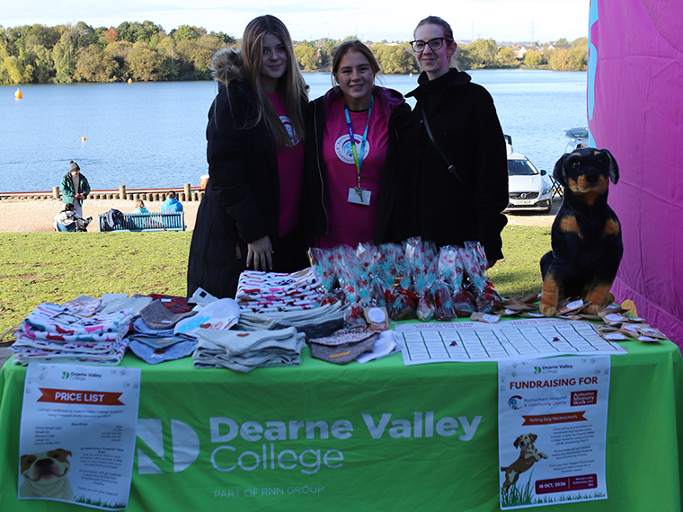 Three people stood at a stall table with a green table cloth, Manvers Lake in the background