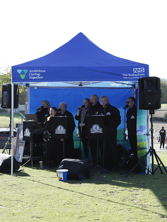 A blue gazebo on grass, several people stood underneath holding microphones singing