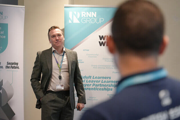 A male in a suit and white shirt stood next to two pull up banners speaking to an audience
