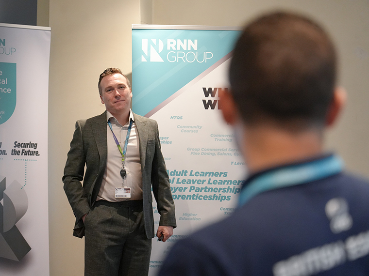 A male in a suit and white shirt stood next to two pull up banners speaking to an audience