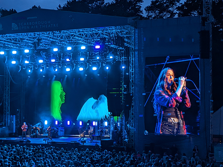 A stage light up at night time and a large screen to the side of it showing a singer, crowds of people stood watching