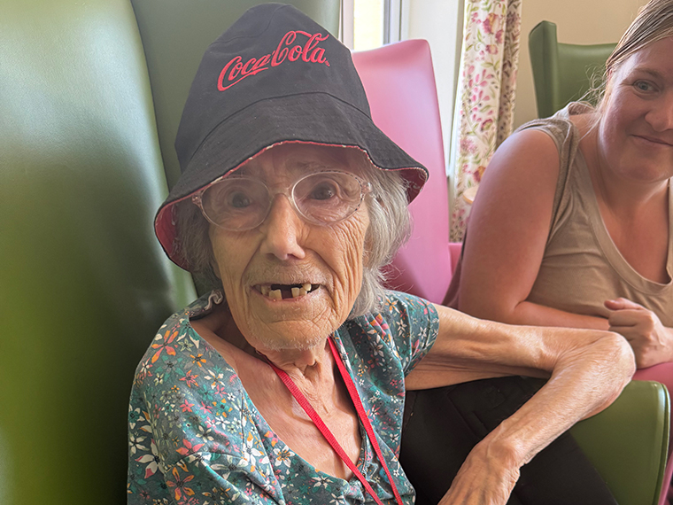 A smiling lady wearing glasses and a black hat with red writing on, sat in a comfy chair