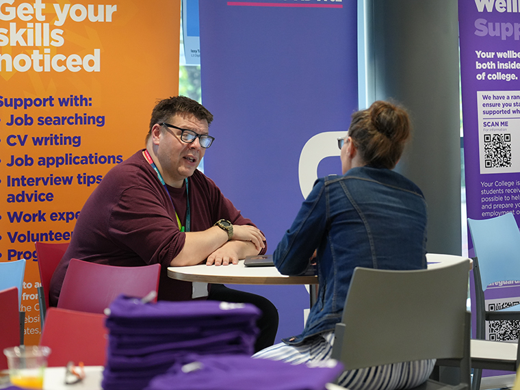 A member of staff talking to a female, both sat around a small round table with pull up banners behind them